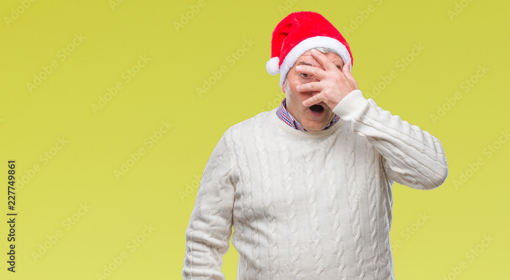 Handsome senior man wearing christmas hat over isolated background peeking in shock covering face and eyes with hand, looking through fingers with embarrassed expression. Handsome senior man wearing christmas hat over isolated background peeking in shock covering face and eyes with hand, looking through fingers with embarrassed expression.