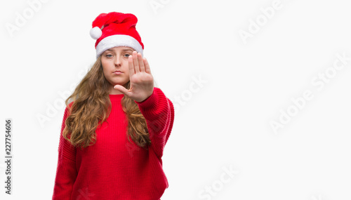 Young blonde woman wearing santa claus hat with open hand doing stop sign with serious and confident expression, defense gesture
