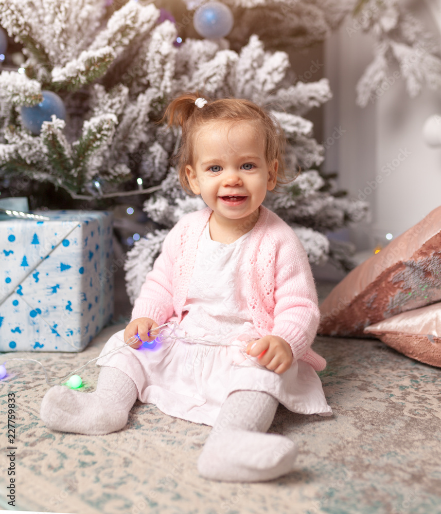 Beautiful cute little girl sitting on the floor near the New Year tree. Holiday mood, childlike spontaneity and sweet atmosphere Beautiful cute little girl sitting on the floor near the New Year tree. Holiday mood, childlike spontaneity and sweet atmosphere