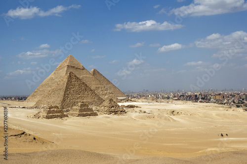 Great Pyramids of Giza set against a bright blue sky and golden yellow desert sands but with the encroaching city of Cairo in the background
