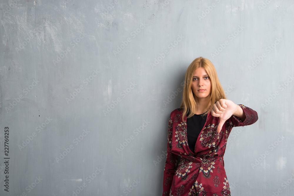 Blonde woman showing thumb down sign with negative expression on textured wall background Blonde woman showing thumb down sign with negative expression on textured wall background