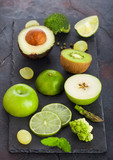 Fresh raw organic green toned fruit and vegetables on stone board black background. Avocado, lime, apple, kiwi and grapes with broccoli and cauliflower. Top view