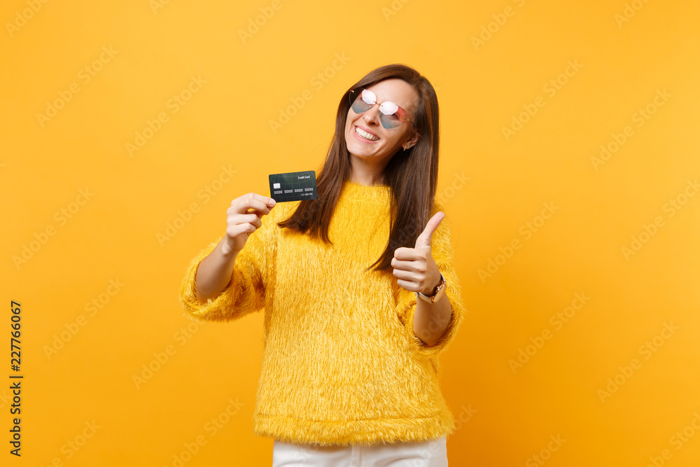 Portrait of joyful young woman in fur sweater, heart eyeglasses showing thumb up holding credit card isolated on bright yellow background. People sincere emotions, lifestyle concept. Advertising area. Portrait of joyful young woman in fur sweater, heart eyeglasses showing thumb up holding credit card isolated on bright yellow background. People sincere emotions, lifestyle concept. Advertising area.