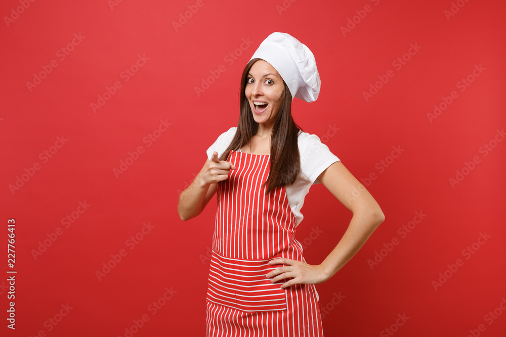 Fototapeta premium Housewife female chef cook or baker in striped apron, white t-shirt, toque chefs hat isolated on red wall background. Beautiful housekeeper woman pointing finger on camera. Mock up copy space concept.