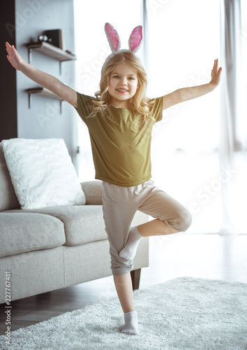Fototapeta Naklejka Na Ścianę i Meble -  I can fly. Full length portrait of carefree small girl standing on one leg while stretching arms sideways. She is laughing. Child is wearing cute bunny ears 