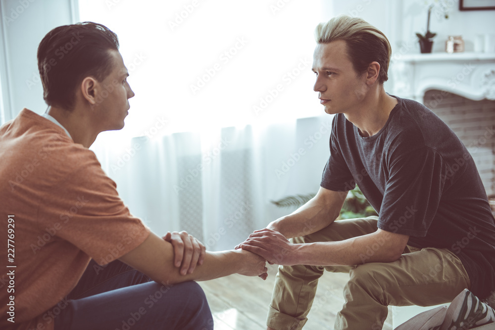 Side view portrait of two handsome guys sitting on couch and looking at each other while sharing tender moment Side view portrait of two handsome guys sitting on couch and looking at each other while sharing tender moment