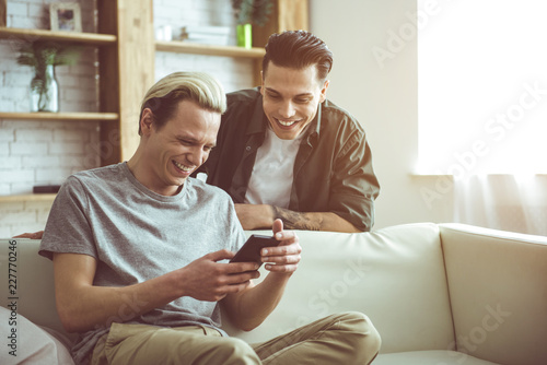 Toned portrait of handsome blond guy sitting on couch and holding mobile phone while his boyfriend standing behind. They looking at cellphone display and laughing