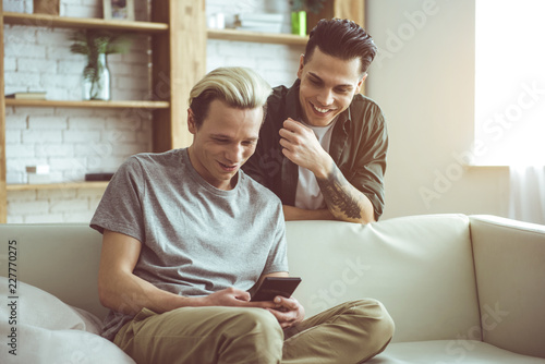 Surfing the internet. Toned portrait of handsome blond guy sitting on couch and holding smartphone while his boyfriend standing behind
