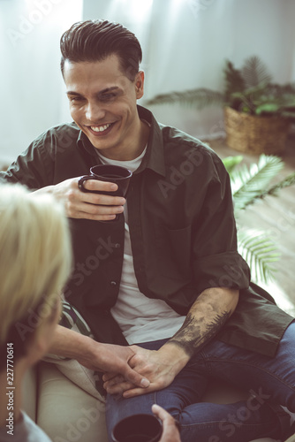 Happy together. Toned portrait of handsome young man with tattoo on arm holding cup of coffee while spending time with friend at home