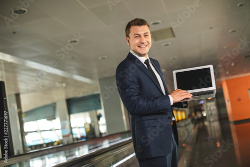 Waist up portrait of blithesome businessman holding laptop and looking at camera with wide smile © Yakobchuk Olena