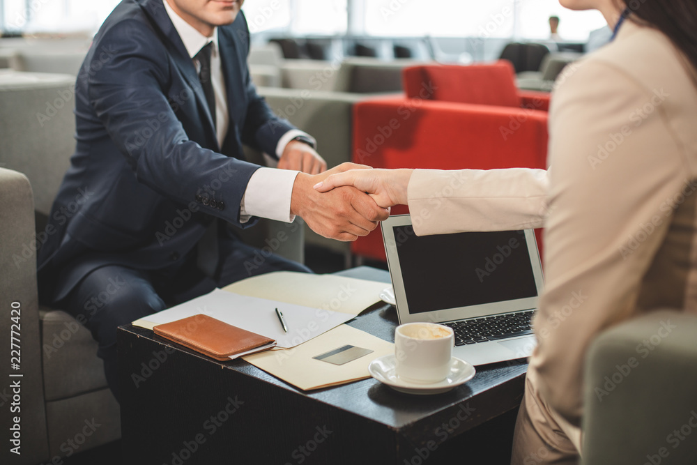 Making a deal. Business partners shaking hands while sitting near table in cafe. Open laptop and papers on counter. Focus on handshake Making a deal. Business partners shaking hands while sitting near table in cafe. Open laptop and papers on counter. Focus on handshake