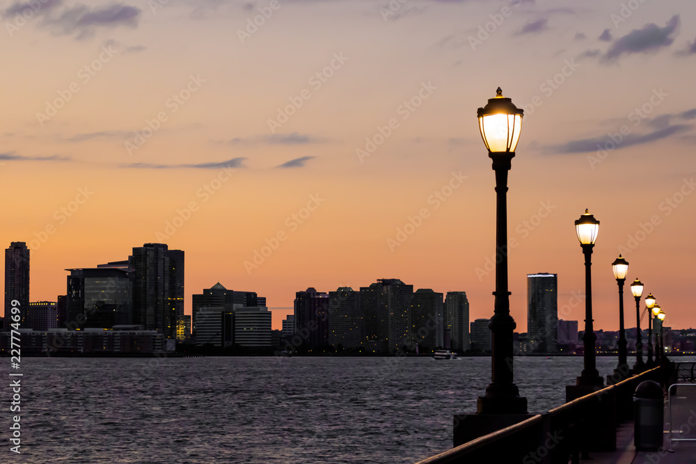 Glowing street lamps at the Finantial District in New York City during sunset with view on Jersey City skyline on the horizon across the Hudson River