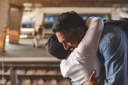 Positive smiling hindu man standing at the railway platform while embracing his little son