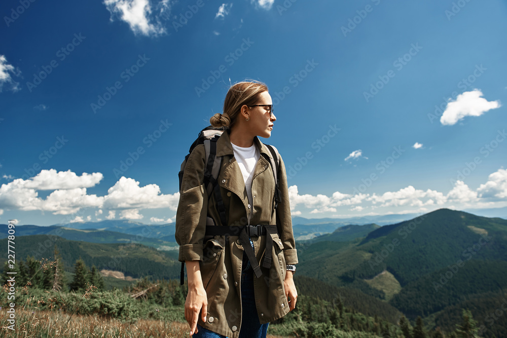 Waist up profile portrait of tranquil lady resting during backpack tour. She standing and looking away observing beautiful landscapes