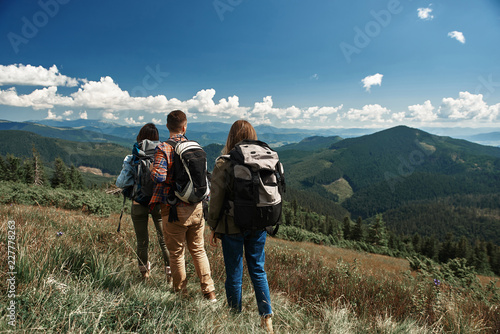 Two women and man carrying rucksacks while tour in highland. They standing on top and looking at valley and peaks in front while focus is on back
