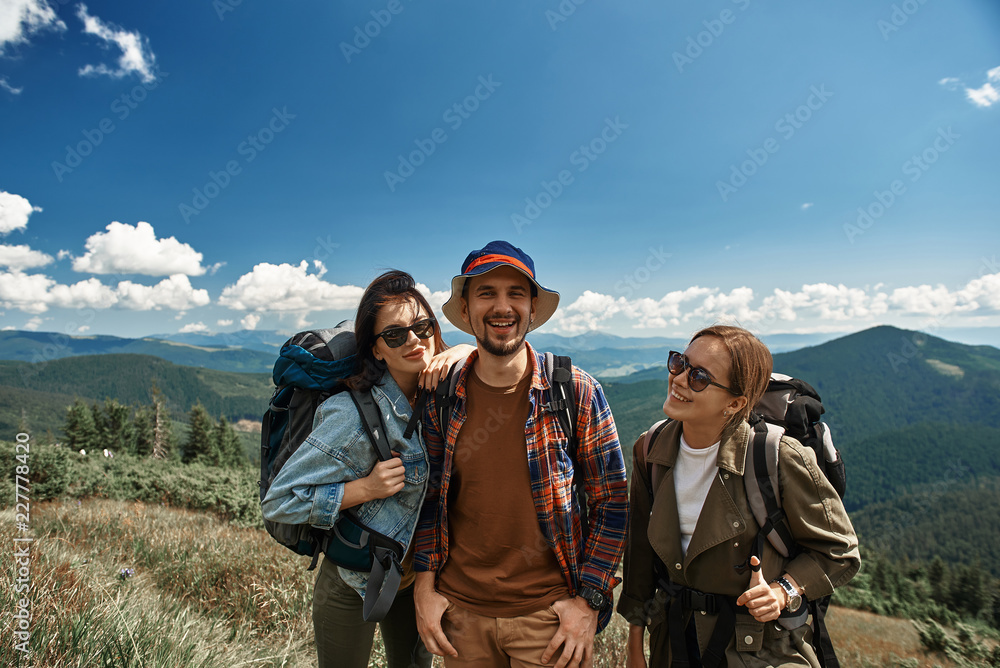 Waist up portrait of two jolly women and man standing in highland. They having fun together on backpack tour among marvelous mountains