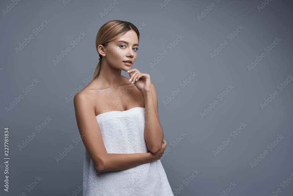 Fototapeta premium Pretty curious lady thoughtfully touching her chin while wearing white towel and standing against the grey background