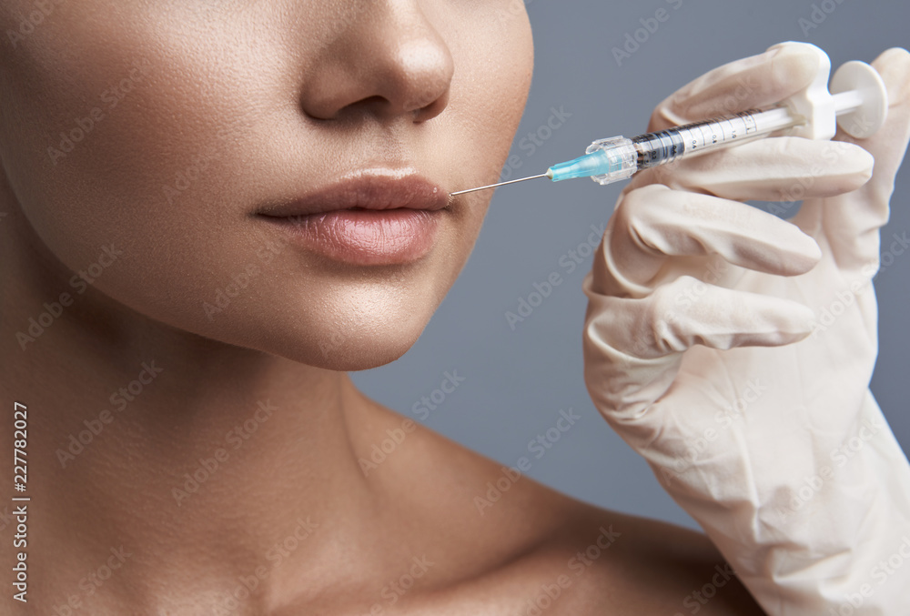 Close up of the young lady standing against the grey background and getting injection in her lip