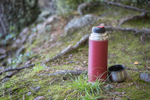 A red thermos and a coffee mug standing in the moss beside a lake in the autumn, outdoor living background