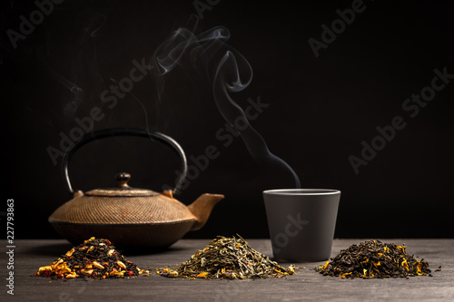 trois petits tas de feuille de thé avec en fond une théière, une tasse et sa fumée