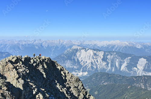 Two climbers at the summit of a steep mountain (Acherkogel, Stubai Alps) near the Inn valley, Tyrol, Austria. Lechtal Alps in the background. Rocky alpine landscape under blue sky.