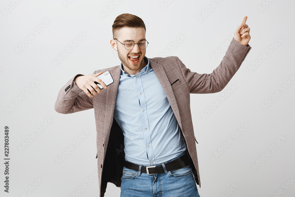 Funny playful and emotive good-looking caucasian guy with bristle and cool haircut dancing joyfully over gray background shaking hands holding smartphone listening music in wireless earphones Funny playful and emotive good-looking caucasian guy with bristle and cool haircut dancing joyfully over gray background shaking hands holding smartphone listening music in wireless earphones