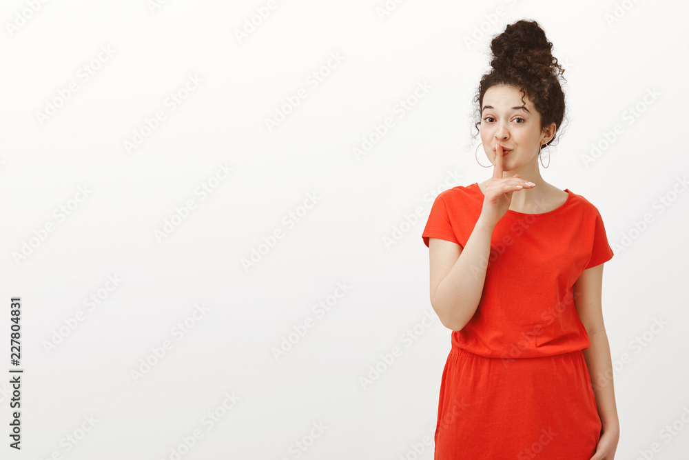 Portrait of creative cheerful Caucasian girlfriend with curly hair combed in bun, smiling curiously while saying shh, making shush gesture with index finger over mouth, making surprise over gray wall Portrait of creative cheerful Caucasian girlfriend with curly hair combed in bun, smiling curiously while saying shh, making shush gesture with index finger over mouth, making surprise over gray wall