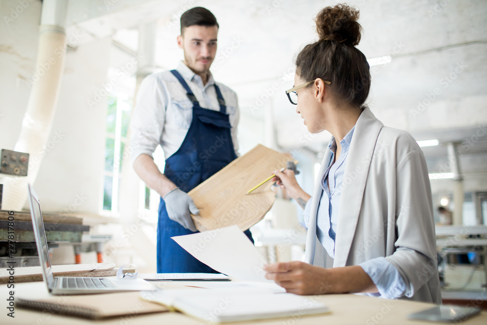 Fototapeta premium Two young contemporary engineers in workwear discussing characteristics of wooden board in workshop