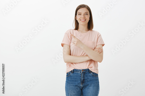 Portrait of charming european female model in pink t-shirt, pointing at upper left corner with index finger and smiling joyfully while standing against gray background, showing new place to friend