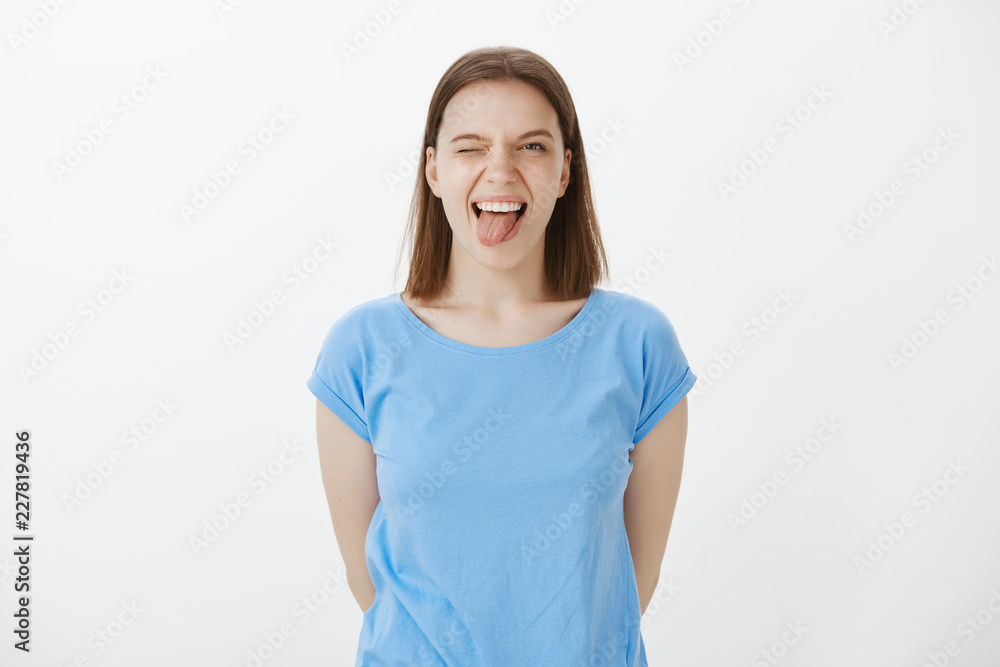 Indoor shot of playful good-looking caucasian woman with fair hair, sticking out tongue and winking, enjoying life and spending great time in friend circle, standing happy and carefree over gray wall Indoor shot of playful good-looking caucasian woman with fair hair, sticking out tongue and winking, enjoying life and spending great time in friend circle, standing happy and carefree over gray wall