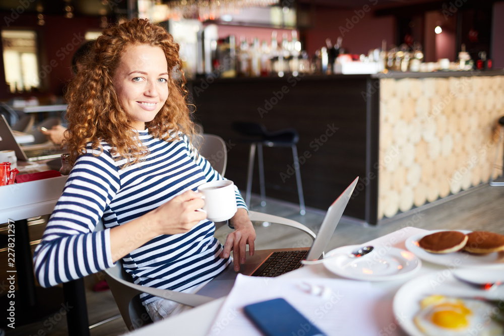 Pretty young cheerful woman with laptop having drink in cafe and networking at break