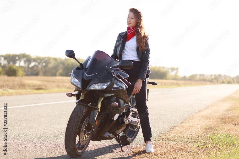 Naklejka premium Transport and extreme concept. Thoughtful stylish female motorcyclist enjoys high speed, poses on fast bike, breathes fresh air, concentrated into distance, wears stylish black clothes for bikers