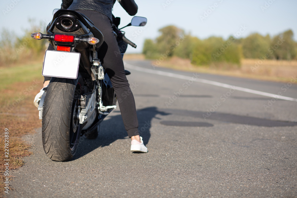 Cropped image of unrecognizable female bikes poses back on bike at asphalt, wears black clothes, blank copy space aside for your advertisement or promotional text. People, transport, destination Cropped image of unrecognizable female bikes poses back on bike at asphalt, wears black clothes, blank copy space aside for your advertisement or promotional text. People, transport, destination