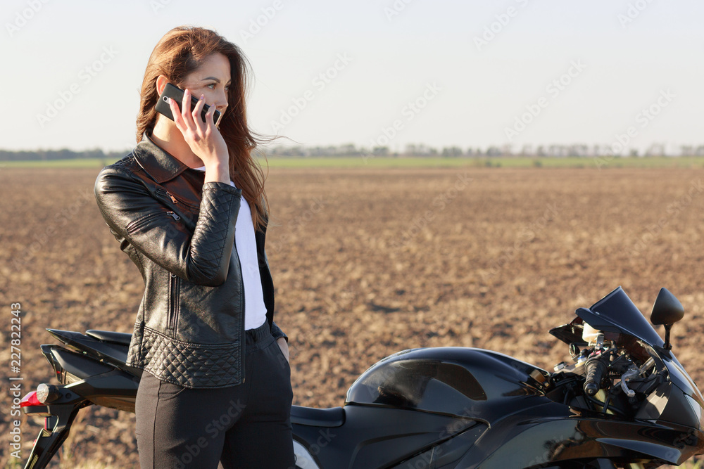 Female biker has phone conversation, stops on road, poses near motorbike, dicusses latest news with friend, dressed in leather jacket and black trousers. Technology and transportation concept Female biker has phone conversation, stops on road, poses near motorbike, dicusses latest news with friend, dressed in leather jacket and black trousers. Technology and transportation concept