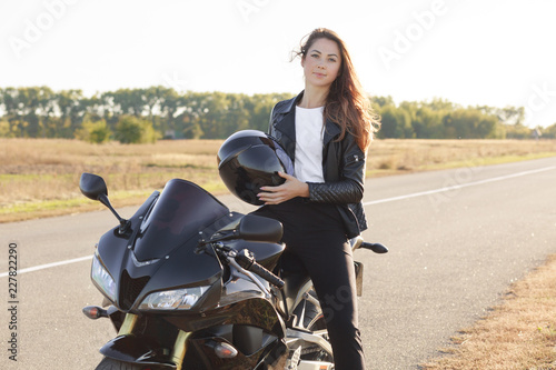 Pleasant looking woman has biker tour  poses on cool bike  holds helmet  wears leather jacket  likes extreme sport  likes driving. People  driving  active lifestyle concept. Biker traveles on morobike
