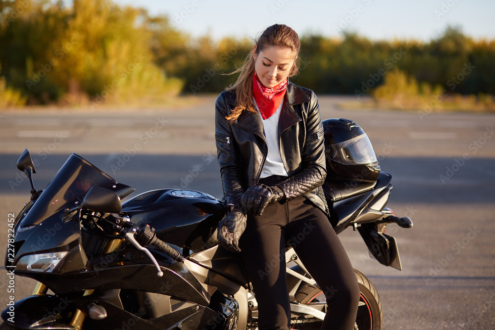 Naklejka premium Outdoor shot of pleased female bikes puts on leather gloves, dressed in black clothes, poses on motorbike, prepares for racing or competitions, poses in countryside. Relaxed woman driver likes hobby
