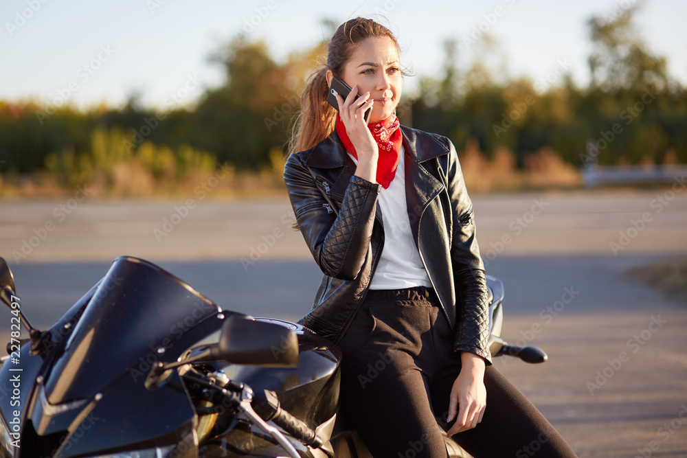 Young female biker with serious expression, has phone conversation via cellular, wears red bandana and leather jacket, poses on motorbike, stops on road to have rest. Technology and lifestyle Young female biker with serious expression, has phone conversation via cellular, wears red bandana and leather jacket, poses on motorbike, stops on road to have rest. Technology and lifestyle