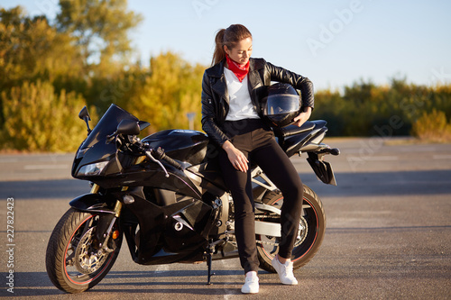 Valokuvatapetti Photo of young female motorcyclist wears fashionable clothes, white sneakers, holds helmet under arm, stands near black motorbike, stands on asphalt, poses outdoor