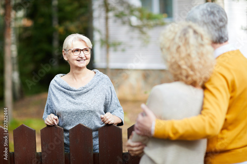 Blonde mature woman in eyeglasses and grey pullover looking at her neighbours during talk through fence photo