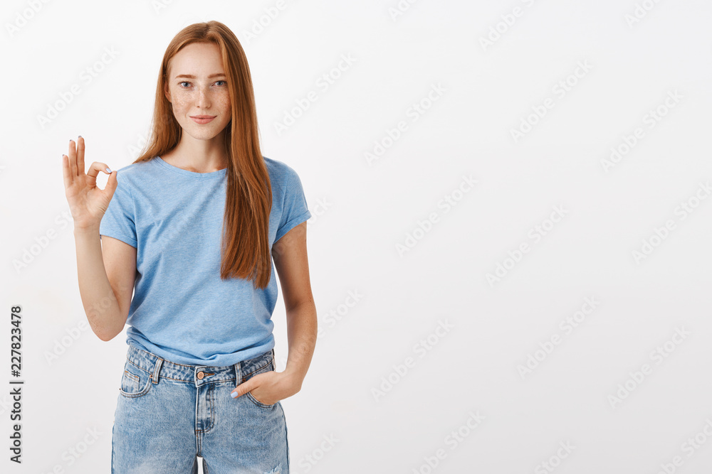 Obraz premium Ok it is deal. Portrait of good-looking satisfied and happy redhead female student in blue t-shirt holding hand in pocket showing okay or perfect gesture with fingers smiling liking or accepting plan