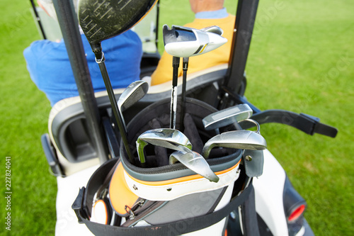 Set of golf clubs in sports bag on back of golf car being driven by one of mature players