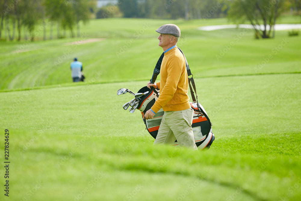 Obraz premium Side view of mature man in casualwear carrying bag with golf clubs while hurrying for game