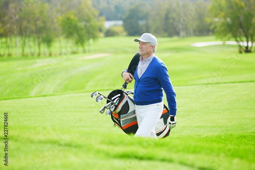 Senior man in casualwear walking for game of golf and carrying clubs in bag