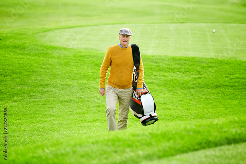 Senior man with sportive bag walking along green field while huttying for game of golf