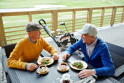 Two friendly senior men sitting in outdoor cafe, having lunch and discussing curious moments of the last game of golf