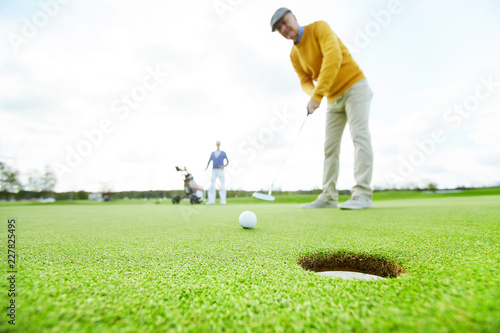 Mature man in casualwear standing on green field and going to hit golf ball into hole