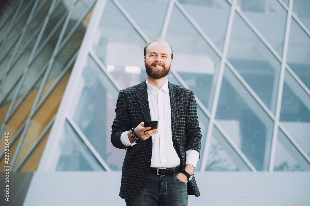 Handsome bearded man smiling and using smartphone outdoor near office Handsome bearded man smiling and using smartphone outdoor near office