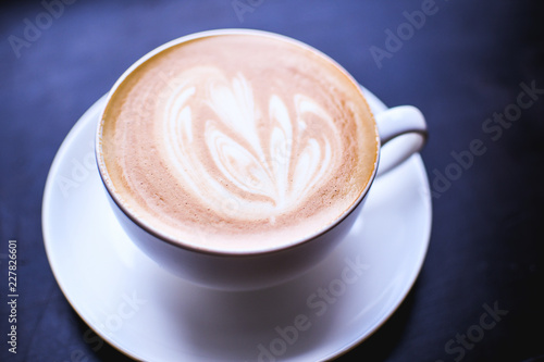 White cup of cappuccino with an abstract drawing on a brown coffee foam. Elegant coffee art. Concept background of cafe, rest, leisure, and romantic settings. Deep black background