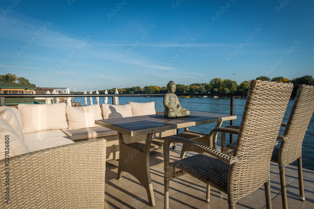 Sofa, chairs and table with Buddha statue on terrace next to woter