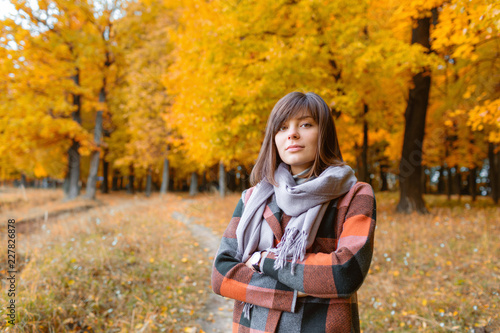 Portrait of young woman in fall forest. Brunette woman in autumn park with fashionable plaid coat and scarf.